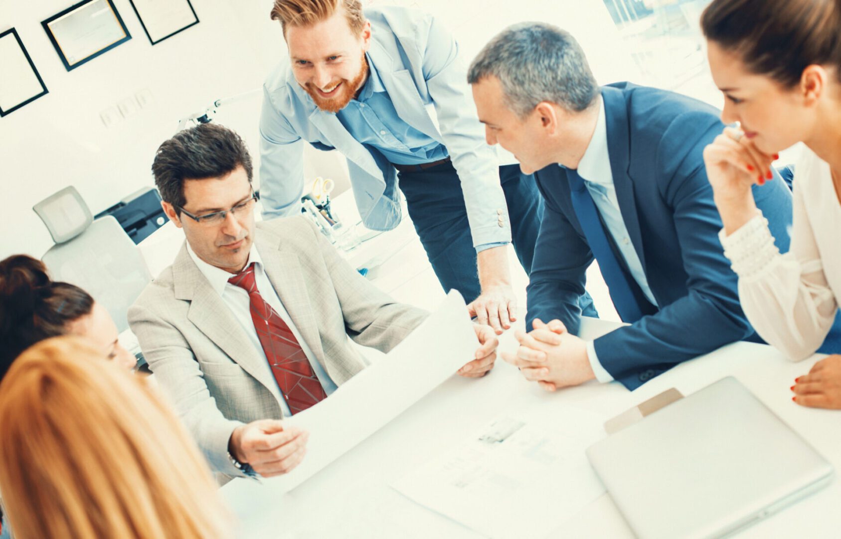 Closeup of mixed age business people in a meeting discussing about the project. One of the mid 40's male coworkers is holding a document which seems to contain positive information. His colleague is leaning on the desk next to her, rest of people are seated. Front view, low angle shot.